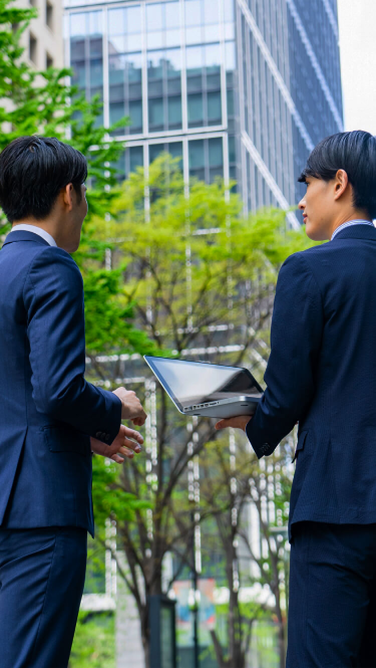 外でパソコンを持った二人のスーツの男性が話している様子